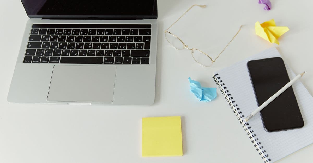 A laptop on an office desk beside a spiral notebook and a crumpled sheet of paper